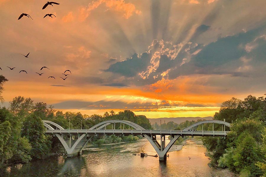 About Our Agency - Bright Orange Sunset with Sun Rays Piercing the Clouds, Birds Flying Past, over Caveman Bridge in Grants Pass, Oregon and the Rogue River