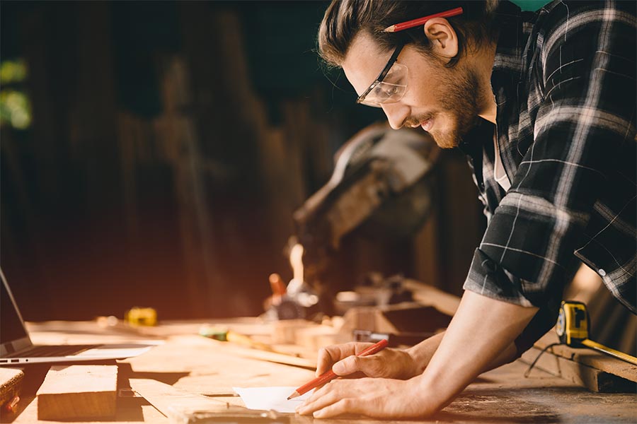 Specialized Business Insurance - Carpenter Marking Measurements, Wearing a Flannel Shirt and Protective Glasses, a Pencil behind His Ear